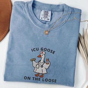 May include: A blue jean Comfort Colors t-shirt with the text "ICU Goose ON THE LOOSE" and an embroidered cartoon goose wearing a doctor's coat. A gold necklace with a pendant is draped across the shirt.