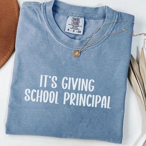 Może przedstawiać: Niebieska koszulka w kolorze dżinsu z białym napisem "IT'S GIVING SCHOOL PRINCIPAL". Złoty naszyjnik z wisiorkiem jest zarzucony na koszulkę. Koszulka jest złożona i ma metkę Comfort Colors.