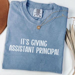 Może przedstawiać: Niebieska koszulka Comfort Colors z napisem "IT'S GIVING ASSISTANT PRINCIPAL" w kolorze białym. Złoty naszyjnik z wisiorkiem jest zarzucony na koszulkę. Koszulka jest złożona i umieszczona na białej powierzchni.