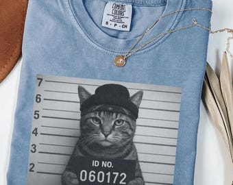 May include: A blue jean coloured t-shirt featuring a cat graphic wearing a hat and holding an ID sign with the number 060172. A gold necklace with a pendant is draped across the shirt.