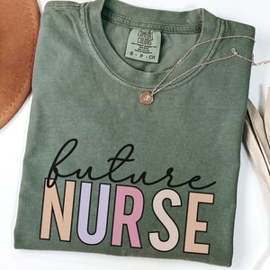 Puede incluir: Una camiseta verde musgo con las palabras "future nurse" impresas en ella. La palabra "nurse" está en letras de bloque de colores.