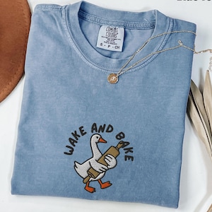 May include: Blue jean Comfort Colors t-shirt with the text "WAKE AND BAKE" and a cartoon goose holding a rolling pin. A gold necklace with a pendant is draped across the shirt. The shirt is size S-P-CH.