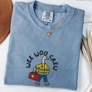Puede incluir: Una camiseta azul vaquera Comfort Colors con el texto "Wee Woo Crew" y un pato de dibujos animados con una chaqueta amarilla y una bolsa médica roja. Un collar dorado con un colgante está sobre la camisa.