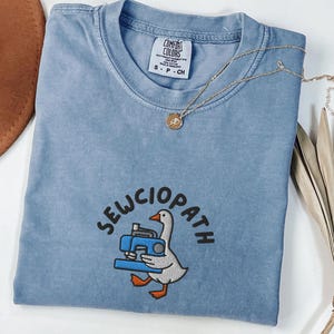 Può includere: Una t-shirt color blu jeans con scollo rotondo ed etichetta Comfort Colors. Il capo presenta un ricamo raffigurante un'oca bianca che tiene una macchina da cucire blu con la scritta SEWCIOPATH arcuata in lettere nere sopra di essa.