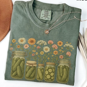 Peut inclure: Un t-shirt vert mousse avec un motif floral et des bocaux de cornichons. Le t-shirt a un col rond et des manches courtes. Un collier argenté avec un petit pendentif est drapé sur le t-shirt. Le mot "Moss" est imprimé en haut à droite.
