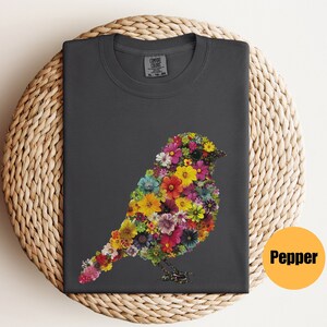 May include: A dark gray t-shirt with a floral bird design. The bird is made up of colorful flowers and is outlined in black. The shirt has a crew neck and short sleeves.
