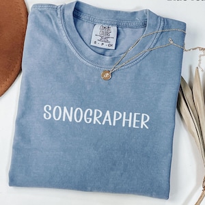 Könnte beinhalten: Ein blaues Jeans-T-Shirt mit dem weißen Aufdruck "SONOGRAPHER". Eine goldene Halskette mit Anhänger liegt über dem Shirt. Das Shirt ist gefaltet und hat ein Etikett mit der Aufschrift "COMFORT COLORS".