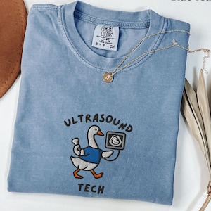 Könnte beinhalten: Hellblaues T-Shirt mit dem Text "ULTRASOUND TECH" und einer Stickerei einer Cartoon-Gans, die ein Ultraschallbild hält. Eine goldene Halskette mit Anhänger liegt über dem Shirt. Das Shirt ist mit "Comfort Colors" gekennzeichnet.