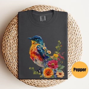 May include: A dark grey t-shirt featuring a colourful bird and flowers printed on the front. The bird is a vibrant mix of blue, yellow, orange, and red. The flowers are a mix of pink, yellow, and blue. The shirt has a crew neck and short sleeves.