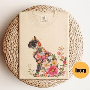 May include: A beige t-shirt featuring a floral cat design. The cat is composed of colourful flowers and is sitting on a white background. The shirt is made from 100% cotton and is available in sizes S-XL. The text "COMFORT COLORS" is printed on the tag.