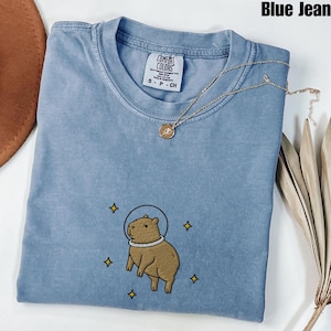 Könnte beinhalten: Ein blaues T-Shirt in Jeansoptik mit einer Stickerei eines braunen Wasserschweins, das einen weißen Weltraumhelm trägt und von kleinen gelben Sternen umgeben ist. Das Shirt hat ein Comfort Colors Markenetikett am Halsausschnitt und eine Goldkette.
