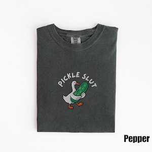 May include: Dark grey t-shirt with a graphic of a white goose holding a green pickle, with the text "PICKLE SLUT" above. The shirt is folded and has a tag that says "Comfort Colors". The word "Pepper" is printed in the bottom right corner.