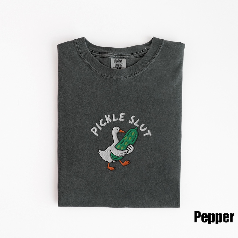 May include: Dark grey t-shirt with a graphic of a white goose holding a green pickle, with the text "PICKLE SLUT" above. The shirt is folded and has a tag that says "Comfort Colors". The word "Pepper" is printed in the bottom right corner.