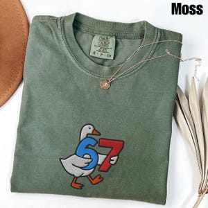 Peut inclure: T-shirt vert mousse Comfort Colors avec un dessin d'oie tenant le chiffre 67. Le t-shirt a un col rond et une petite &eacute;tiquette. Un collier en or avec un pendentif est drap&eacute; sur le t-shirt.