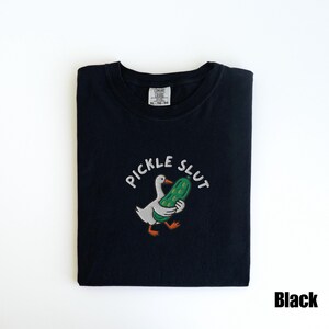 May include: Black t-shirt with the embroidered phrase "PICKLE SLUT" above a cartoon duck holding a green pickle. The shirt is folded and has a tag in the upper center.