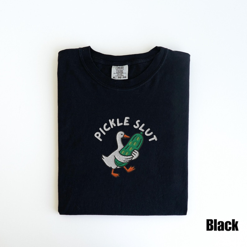 May include: Black t-shirt with the embroidered phrase "PICKLE SLUT" above a cartoon duck holding a green pickle. The shirt is folded and has a tag in the upper center.