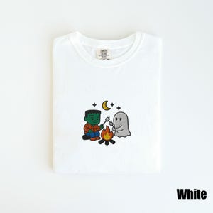 May include: White t-shirt with an embroidered Halloween design. The design features a Frankenstein monster roasting marshmallows with a ghost over a campfire, under a crescent moon and stars. The word "White" is printed at the bottom.