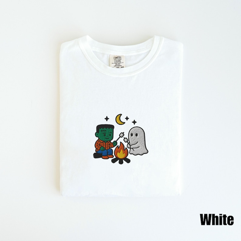 May include: White t-shirt with an embroidered Halloween design. The design features a Frankenstein monster roasting marshmallows with a ghost over a campfire, under a crescent moon and stars. The word "White" is printed at the bottom.