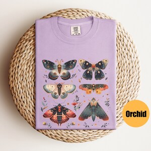Puede incluir: Una camiseta de color lavanda con un estampado de seis polillas coloridas con detalles intrincados. Las polillas est&aacute;n dispuestas en un patr&oacute;n sim&eacute;trico en la camiseta. La camiseta tiene una etiqueta que dice "Comfort Colors" y "XL - 75-80".