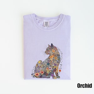 May include: A lavender t-shirt with a floral cat design. The cat is made up of colorful flowers and is sitting on a light purple background. The shirt has a tag that says "Comfort Colors" and "XL - Y/S - 60".