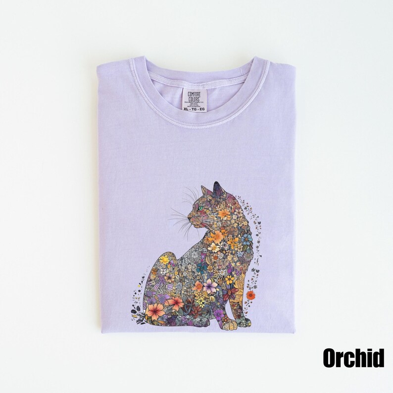 May include: A lavender t-shirt with a floral cat design. The cat is made up of colorful flowers and is sitting on a light purple background. The shirt has a tag that says "Comfort Colors" and "XL - Y/S - 60".