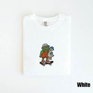 May include: White t-shirt with a colorful embroidered design. The design features a green frog wearing a hat and orange overalls, holding a white duck, both on a skateboard. The text "White" is at the bottom right.