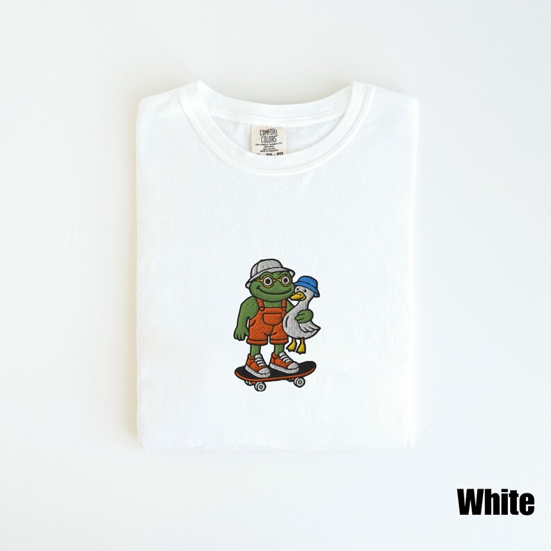 May include: White t-shirt with a colorful embroidered design. The design features a green frog wearing a hat and orange overalls, holding a white duck, both on a skateboard. The text "White" is at the bottom right.