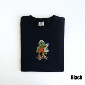May include: Black t-shirt featuring an embroidered design of a frog and a duck on a skateboard. The frog wears glasses, a white hat, and orange overalls. The duck wears a blue hat. The shirt has the text "Black".