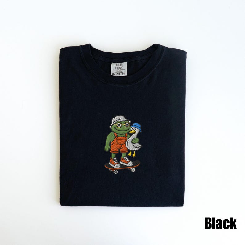May include: Black t-shirt featuring an embroidered design of a frog and a duck on a skateboard. The frog wears glasses, a white hat, and orange overalls. The duck wears a blue hat. The shirt has the text "Black".