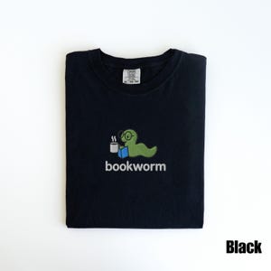 May include: Black t-shirt with a green bookworm graphic wearing glasses and holding a coffee cup. The word "bookworm" is embroidered below the graphic. The shirt is folded and has a tag in the upper center.