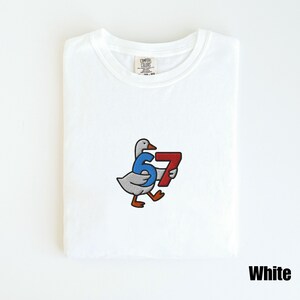 Peut inclure: T-shirt blanc avec une oie de dessin anim&eacute; tenant les chiffres 6 et 7. Les chiffres sont bleus et rouges. Le t-shirt a un col rond et le mot "White" est imprim&eacute; en bas &agrave; droite.