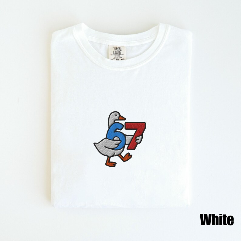Peut inclure: T-shirt blanc avec une oie de dessin anim&eacute; tenant les chiffres 6 et 7. Les chiffres sont bleus et rouges. Le t-shirt a un col rond et le mot "White" est imprim&eacute; en bas &agrave; droite.