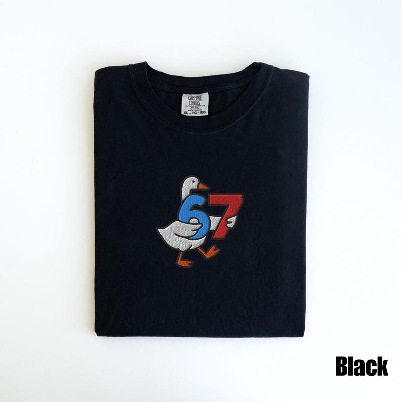 Peut inclure: T-shirt noir avec un motif brod&eacute; d'une oie tenant les chiffres 6 et 7 en bleu et rouge. Le t-shirt est pli&eacute; avec une &eacute;tiquette en haut. Le mot "Black" est dans le coin inf&eacute;rieur droit.