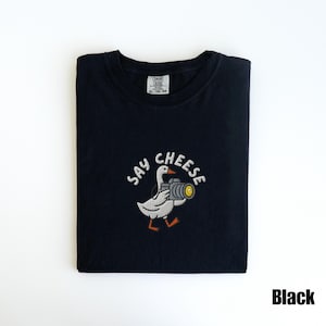 May include: Black t-shirt with a white embroidered design featuring a goose holding a camera, with the words "SAY CHEESE" above. The shirt is folded and has a tag in the upper center.