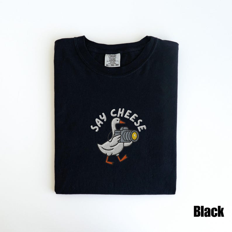 May include: Black t-shirt with a white embroidered design featuring a goose holding a camera, with the words "SAY CHEESE" above. The shirt is folded and has a tag in the upper center.