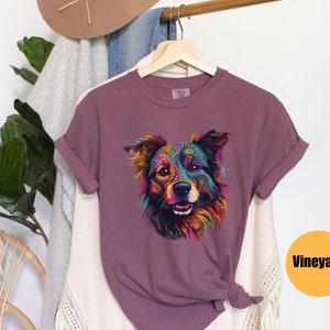 May include: A mauve t-shirt featuring a colourful graphic of a dog's face. The dog has a black nose and brown eyes. The graphic is surrounded by a white border. The t-shirt is hanging on a hanger. The word "Vineyard" is printed in black on a yellow circle.