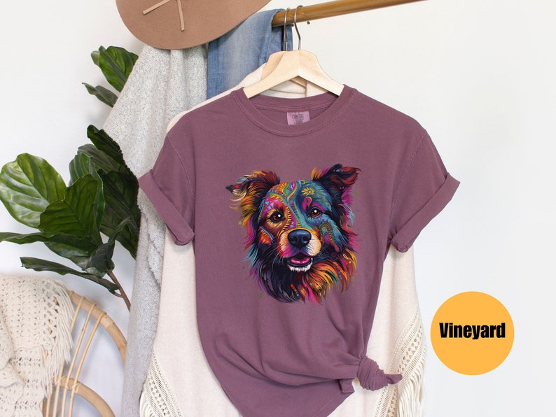 May include: A mauve t-shirt featuring a colourful graphic of a dog's face. The dog has a black nose and brown eyes. The graphic is surrounded by a white border. The t-shirt is hanging on a hanger. The word "Vineyard" is printed in black on a yellow circle.