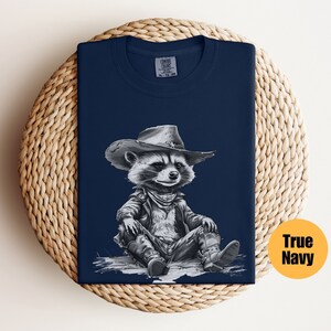 May include: A navy blue t-shirt featuring a black and white illustration of a raccoon wearing a cowboy hat and a leather jacket. The raccoon is sitting with its legs crossed and wearing cowboy boots.