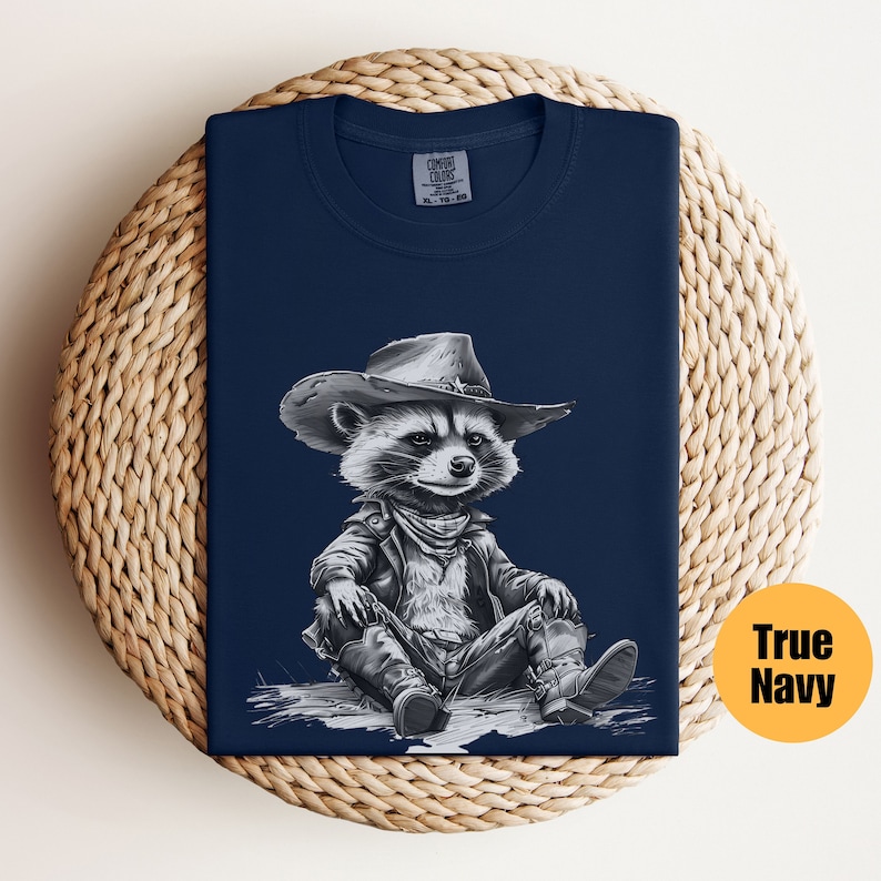 May include: A navy blue t-shirt featuring a black and white illustration of a raccoon wearing a cowboy hat and a leather jacket. The raccoon is sitting with its legs crossed and wearing cowboy boots.