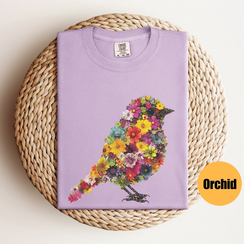 May include: A lavender t-shirt with a floral bird design. The bird is made up of colorful flowers and is outlined in black. The shirt has a crew neck and short sleeves.