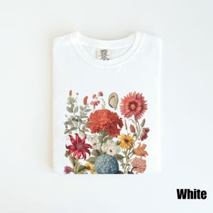 Pu&ograve; includere: Maglietta bianca con stampa floreale che presenta una variet&agrave; di fiori in tonalit&agrave; di rosso, rosa, arancio, giallo e blu. I fiori sono disposti in un design a bouquet e sono posizionati su uno sfondo bianco.