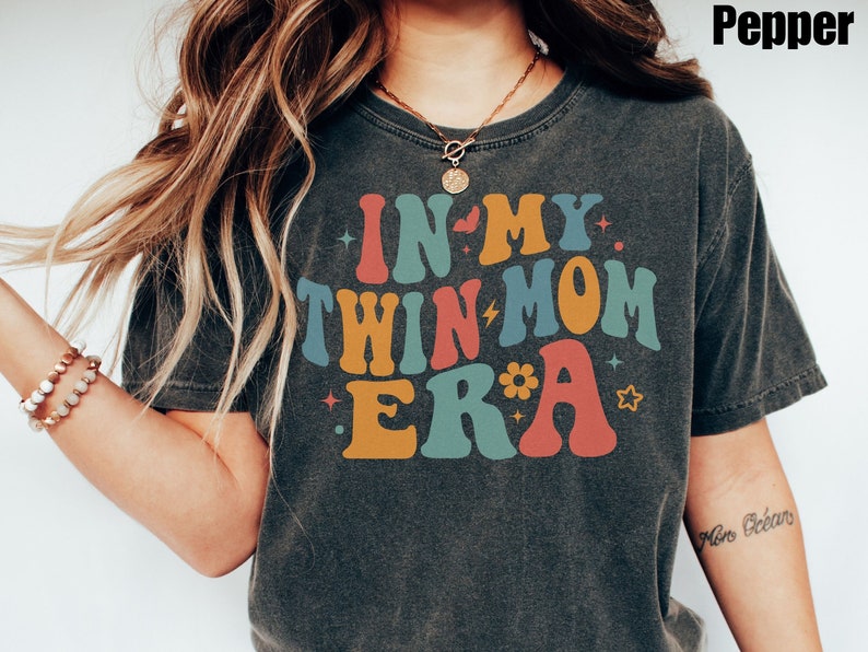 Może przedstawiać: Szary t-shirt z napisem "In My Twin Mom Era" w kolorowych, retro literach. Tekst jest otoczony gwiazdami i kwiatem.