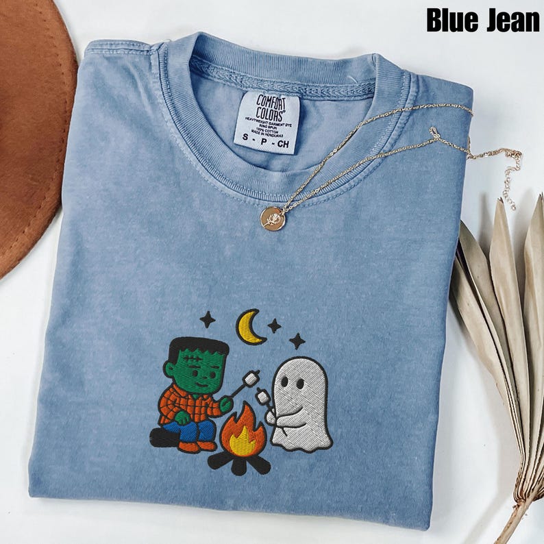 May include: A blue Comfort Colors t-shirt with an embroidered design of a Frankenstein-like character and a ghost roasting marshmallows over a campfire. The shirt has a crew neck and short sleeves. A gold necklace with a pendant is draped across the shirt. The text "Blue Jean" is at the top.