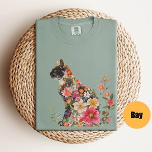 May include: A light green t-shirt featuring a floral cat design. The cat is made up of colourful flowers and leaves. The shirt has a crew neck and short sleeves.