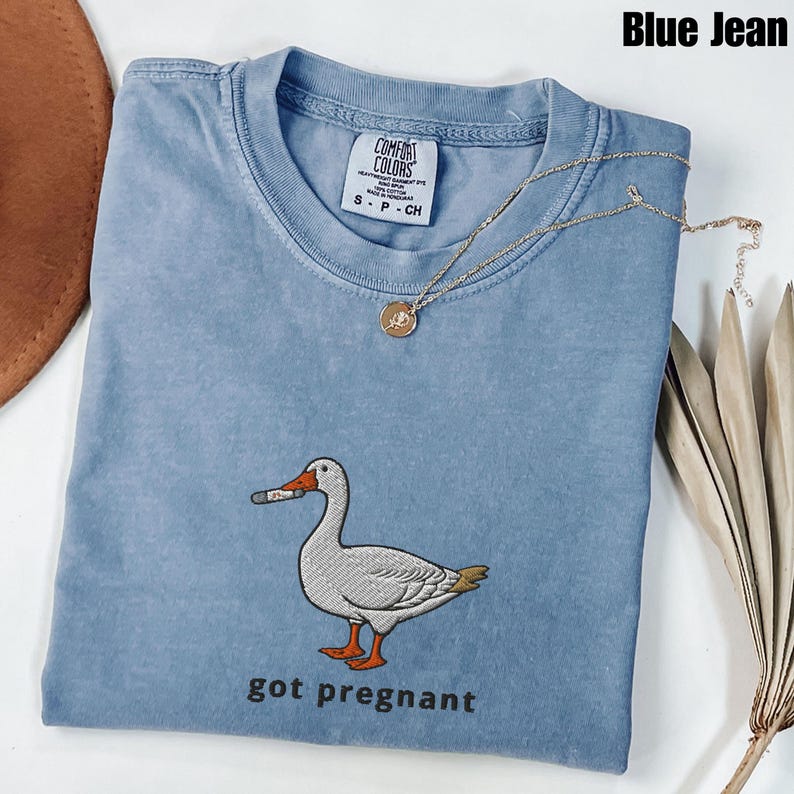 May include: A blue jean Comfort Colors t-shirt with an embroidered goose smoking a cigarette, with the text "got pregnant" below the goose. A gold necklace with a pendant is draped across the shirt.