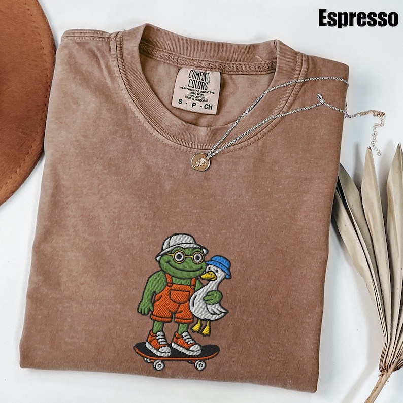 May include: Espresso-colored Comfort Colors t-shirt with an embroidered frog and duck design. The frog wears a hat, glasses, and orange overalls, holding a duck on a skateboard. A silver necklace with a pendant is draped across the shirt.