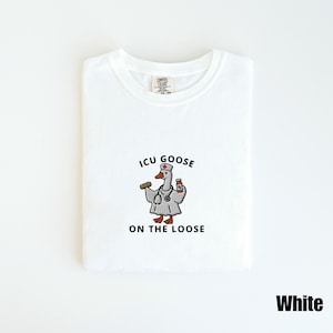 May include: White t-shirt with a crew neck. The shirt features a cartoon goose wearing a doctor's coat and holding a syringe and medicine bottle. The text "ICU GOOSE ON THE LOOSE" is printed below the goose.