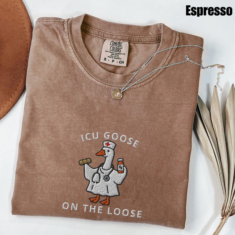 May include: Espresso-colored t-shirt with the text "ICU GOOSE ON THE LOOSE" and an embroidered cartoon goose wearing a nurse's uniform. The shirt has a crew neck and short sleeves. A silver necklace with a small pendant is draped across the shirt.