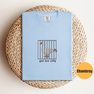 May include: A light blue t-shirt with a white goose in a cage and the text "got too silly". The t-shirt is made of Chambray fabric.