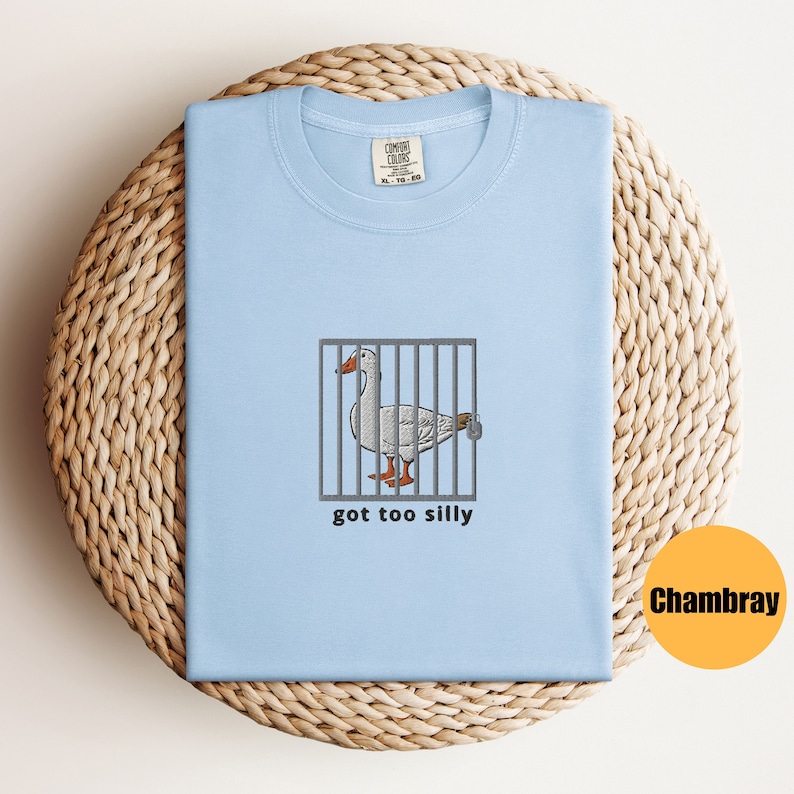 May include: A light blue t-shirt with a white goose in a cage and the text "got too silly". The t-shirt is made of Chambray fabric.